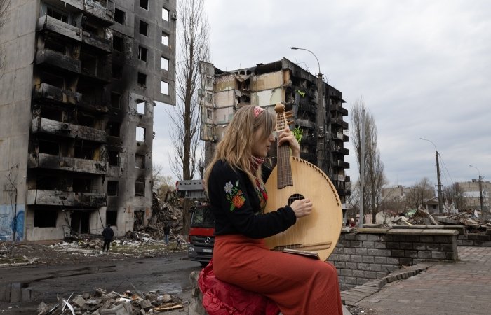 A woman plays a stringed instrument in front of a destroyed building in Bodoryanca, Kyiv Oblast, Ukraine