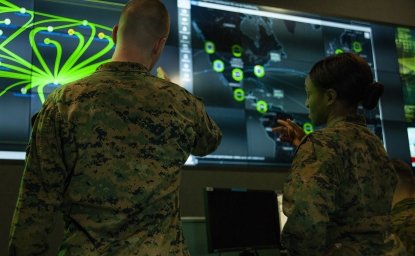 Two soldiers have their back to the camera while looking at a green and blue display of digital information.